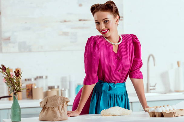 smiling pin up girl in crimson dress and blue apron standing by kitchen table - 写真・画像