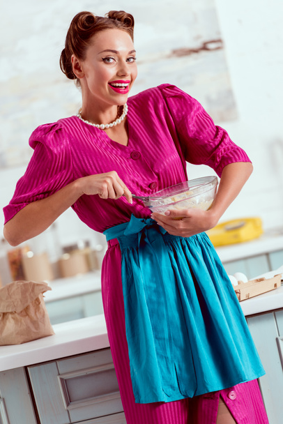 Smiling pin up girl in crimson dress and blue apron mixing ingredients for dough - 写真・画像