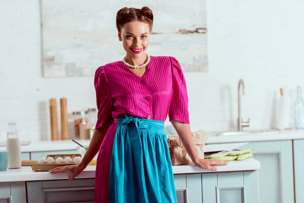 Beautiful pin up girl standing in light blue kitchen near table with different products and looking at camera - Foto, afbeelding
