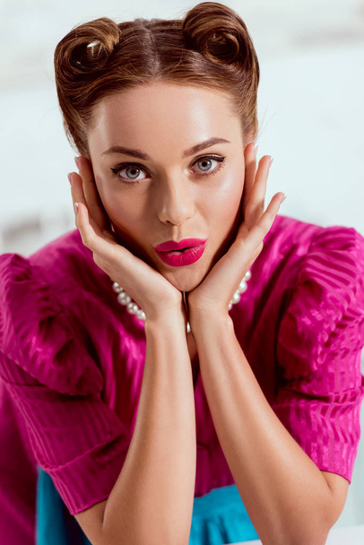 Beautiful pin up girl in crimson dress and pearl necklace holding head in hands and looking at camera - Фото, изображение