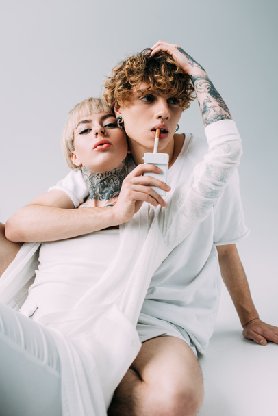man with curly hair holding cigarette in mouth near blonde tattooed girl isolated on grey - Фото, изображение
