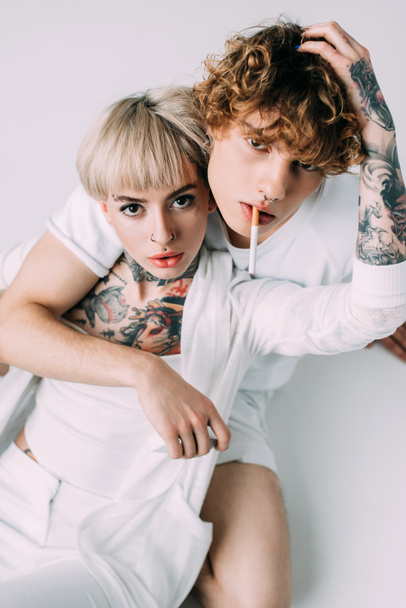 handsome man with curly hair holding cigarette in mouth while sitting with blonde tattooed girl isolated on grey - Foto, Imagen