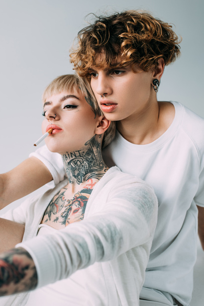 blonde tattooed girl holding cigarette in mouth near handsome man with curly hair isolated on grey  - Photo, Image
