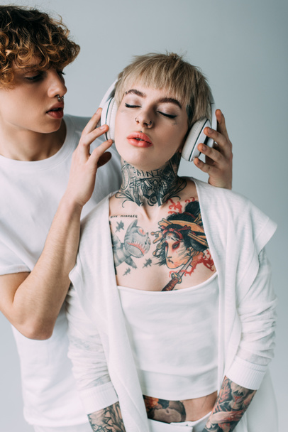 クローズドの刺青の女の子のヘッドフォンを保持している巻き毛の男目グレーに分離された音楽を聴く  - 写真・画像