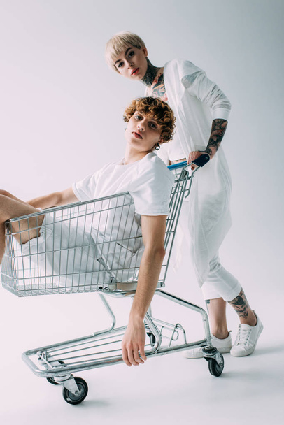 attractive blonde girlfriend pushing shopping cart with boyfriend inside isolated on grey - Фото, изображение