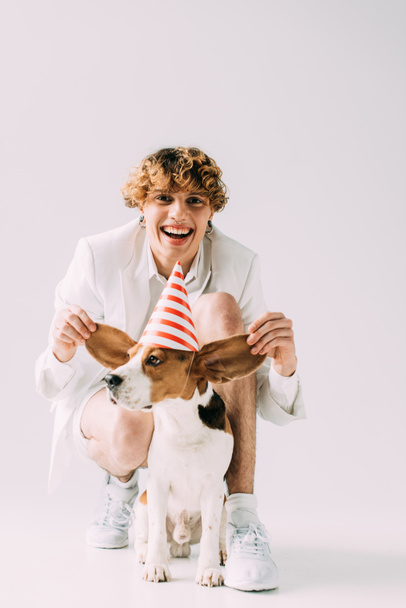 happy man with curly hair holding ears of cute beagle dog on grey background - Photo, Image