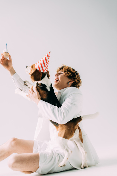 glücklicher Mann mit lockigem Haar, der Cupcake über dem Kopf in der Nähe von Beagle-Hund in Partymütze auf grauem Hintergrund hält - Foto, Bild