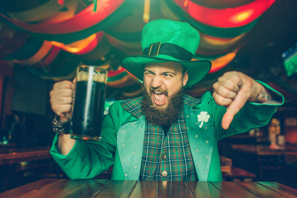Upset and disgussed young man in green suit pose on camera. He sit at table in pub and show big thumb down. Young man hold mug of dark beer. - Foto, immagini