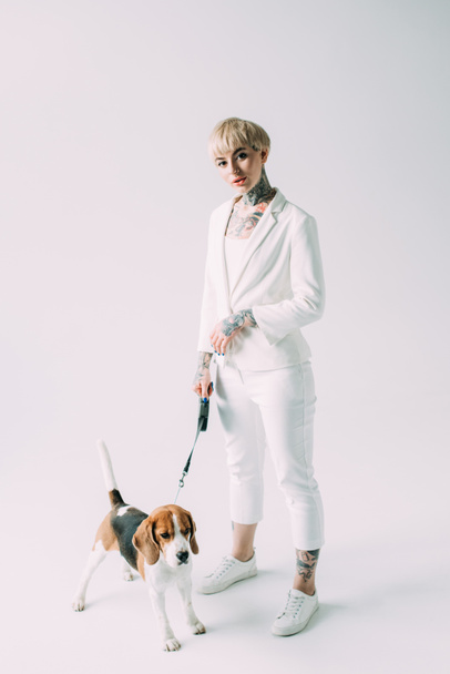 attractive blonde woman holding leash while standing with beagle dog on grey background - Fotó, kép