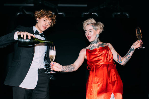 knappe man met krullend haar gieten van champagne in glas blonde vrouw in rode jurk op zwarte achtergrond - Foto, afbeelding