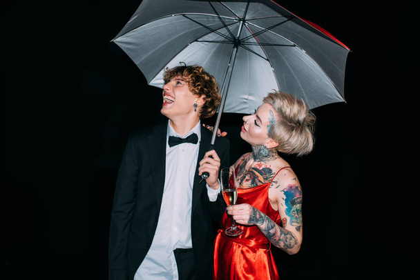 fröhliches Paar, das unter einem Regenschirm steht und isoliert auf schwarz lächelt - Foto, Bild