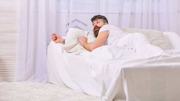 Man in shirt laying on bed awake, white curtain on background. Wake up and oversleep concept. Macho with beard and mustache overslept waking up call. Guy on surprised face waking up in morning - Photo, image