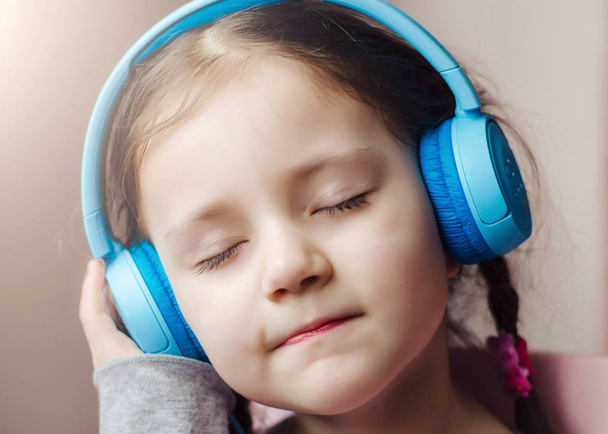 Little girl with blue headphones, holding a smart phone in hands and sitting in bed. Little girl dressed in pink pajamas. Selective focus. Girl is listening music with closed eyes. - Photo, Image