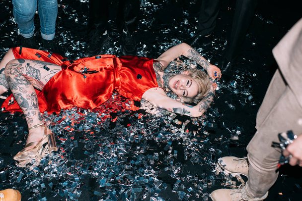 szőke nő feküdt padlón, konfetti közelében emberek a fekete háttér - Fotó, kép