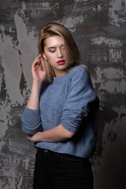 Lovely blonde woman with lush hair wearing blue sweater, posing in studio with shadows - Foto, afbeelding
