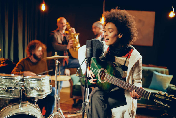 Mixed race woman singing and playing guitar while sitting on chair with legs crossed. In background drummer, saxophonist and bass guitarist. - Photo, Image