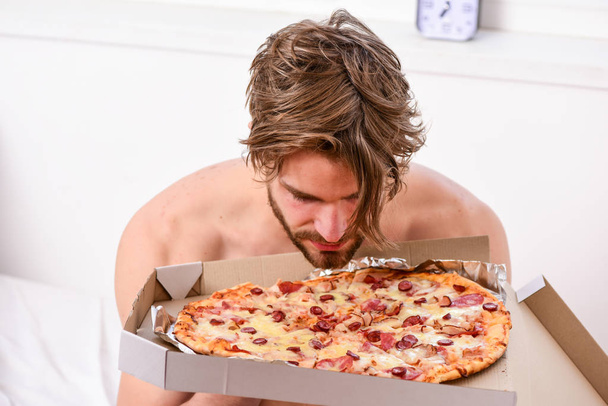 Essenslieferservice. Porträt eines faulen, muskulösen Mannes, der Pizza isst, während er zu Hause auf einem Bett liegt. Mann bärtigen gutaussehenden Kerl essen kitschiges Essen zum Frühstück im Bett. Pizzabett. - Foto, Bild
