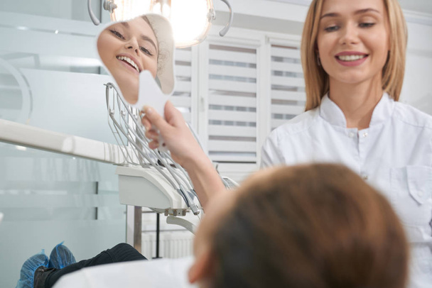 Жінка в стоматологічному кріслі дивиться на дзеркало, посміхаючись
. - Фото, зображення