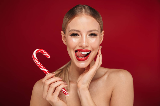Piękno portret atrakcyjna blond włosach topless kobieta stojąc na białym tle nad czerwonym tle, jedzenie cukierków Boże Narodzenie - Zdjęcie, obraz