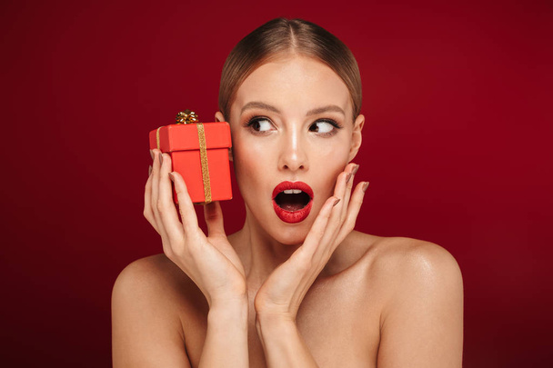 Beauty portrait of an attractive blonde haired topless woman standing isolated over red background, holding gift box - Fotografie, Obrázek