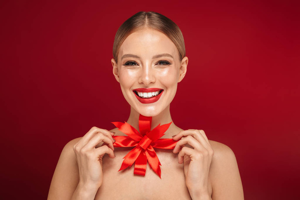 Beauty portrait of a pretty young topless woman standing isolated over red background, holding Christmas bow - Fotoğraf, Görsel