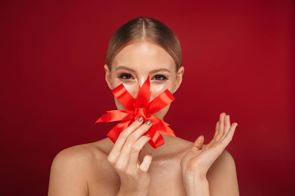 Beauty portrait of an attractive blonde haired topless woman standing isolated over red background, holding red bow - 写真・画像