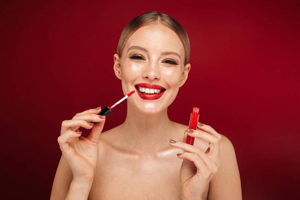 Beauty portrait of a pretty young topless woman wearing makeup isolated over red background, applying lip gloss - Фото, изображение