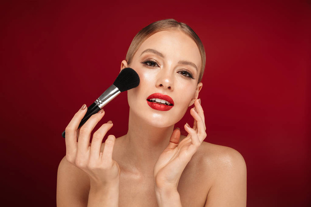 Beauty portrait of a pretty young topless woman wearing makeup isolated over red background, applying makeup with a brush - Fotografie, Obrázek
