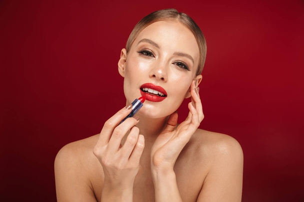 Beauty portrait of a pretty young topless woman wearing makeup isolated over red background, applying red lipstick - Fotó, kép