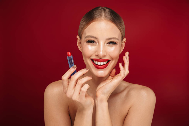 Beauty portrait of a pretty young topless woman wearing makeup isolated over red background, applying red lipstick - Φωτογραφία, εικόνα