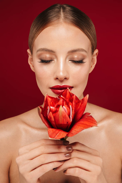 Beauty portrait of an attractive blonde haired topless woman standing isolated over red background, posing with red flower - Photo, image