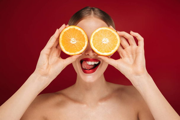 Beauty portrait of a pretty young topless woman wearing makeup isolated over red background,showing sliced orange - Photo, image