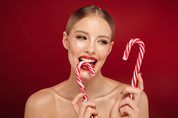 Beauty portrait of a pretty young topless woman wearing makeup isolated over red background, holding christmas candy cone - Foto, Imagen