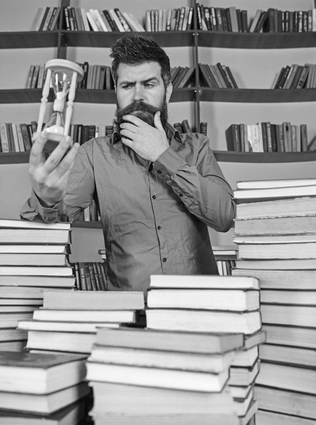 Librarian concept. Man on thoughtful face stands between piles of books, while holds hourglass, bookshelves on background. Teacher, scientist with beard stands at table with books, defocused - Foto, immagini