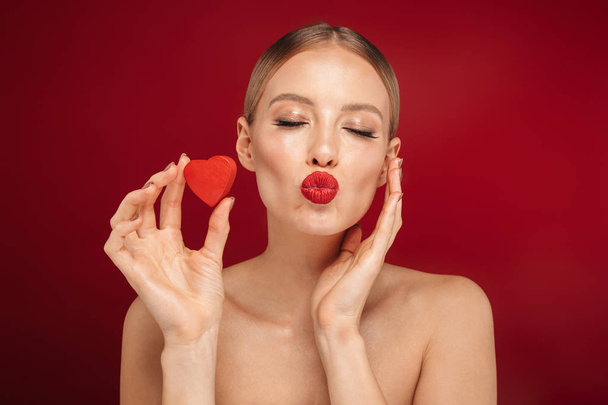 Beauty portrait of an attractive blonde haired topless woman standing isolated over red background, showing little heart - 写真・画像