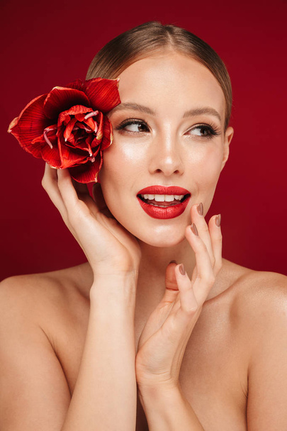 Beauty portrait of an attractive blonde haired topless woman standing isolated over red background, posing with red flower - Fotó, kép