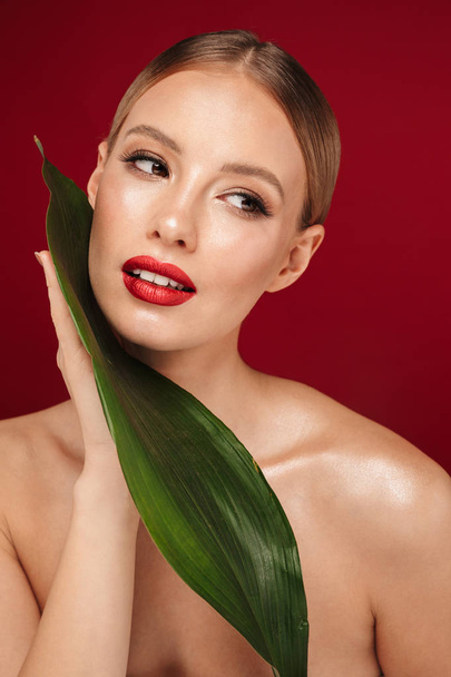 Beauty portrait of an attractive blonde haired topless woman standing isolated over red background, posing with green leaf - Φωτογραφία, εικόνα