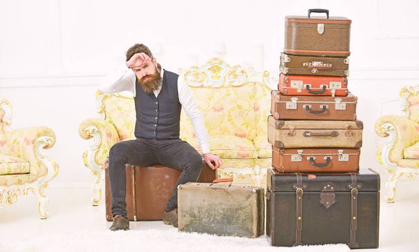 Macho elegant on strict face sits tired near pile of vintage suitcase. Man, butler with beard and mustache delivers luggage, luxury white interior background. Luggage and relocation concept - Zdjęcie, obraz