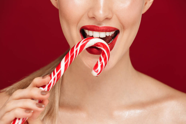 Cropped portrait of a pretty young topless woman standing isolated over red background, eating Christmas candy cone - 写真・画像
