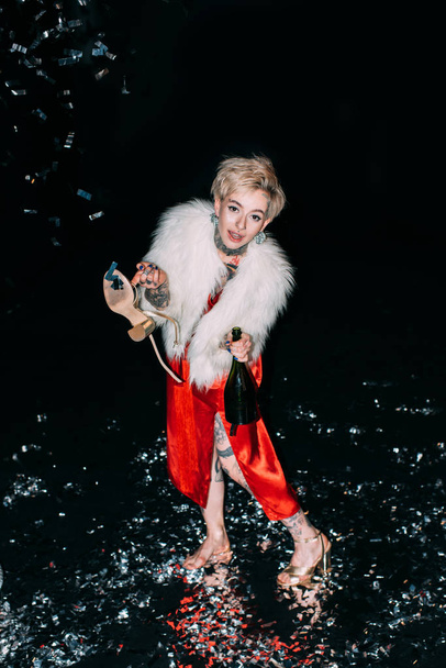 blonde woman with tattoos holding bottle of champagne and shoe isolated on black - Fotó, kép