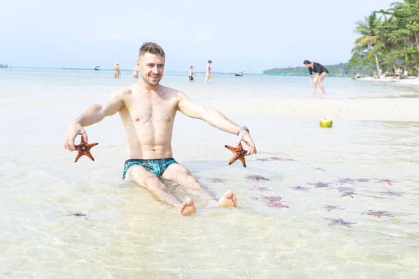 Travel boy is sitting on the sea with red starfishes makes funny photos in Phu Quoc island in crystal clear water - Foto, afbeelding