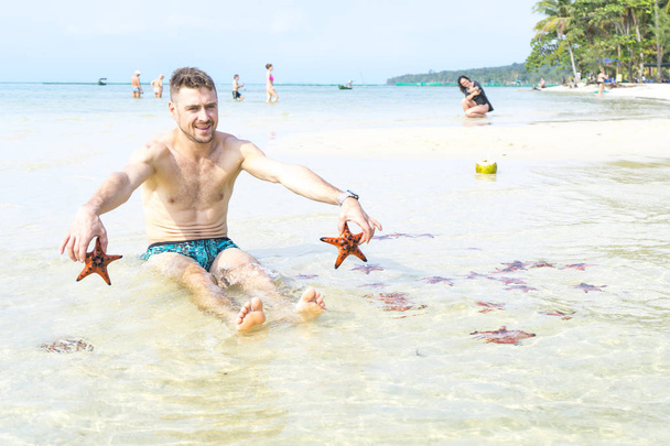 Travel boy is sitting on the sea with red starfishes makes funny photos in Phu Quoc island in crystal clear water - Foto, imagen