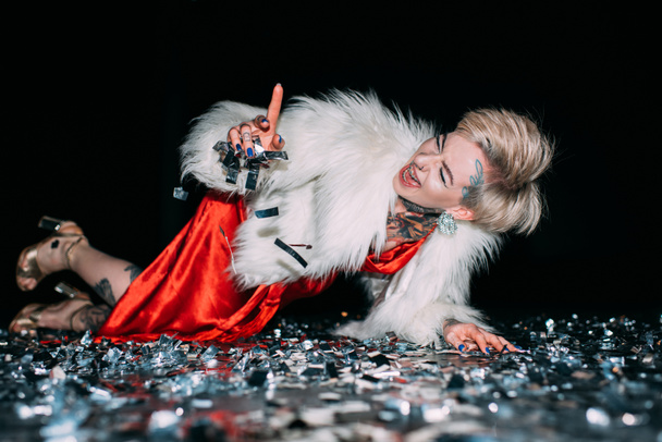 cheerful blonde woman pointing with finger while lying in floor with confetti isolated on black - 写真・画像