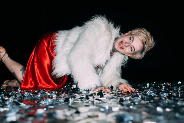 cheerful blonde woman with tattoos lying in floor with confetti isolated on black - Φωτογραφία, εικόνα