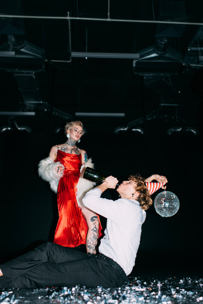 blonde woman looking at man holding disco ball and drinking from bottle on black background  - Fotó, kép