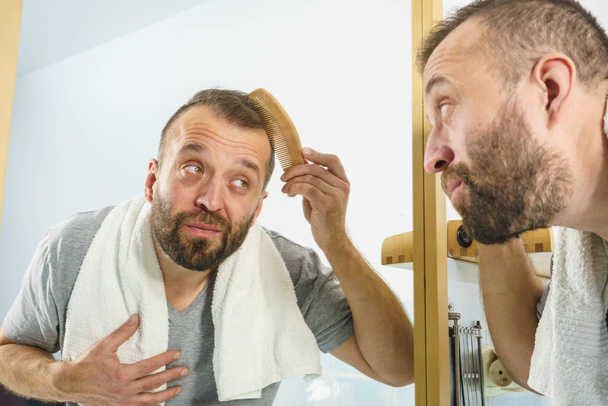 Adult man standing in front of the bathroom mirror brushing his short hair using comb. Guy investigating his receding hairline - Фото, изображение