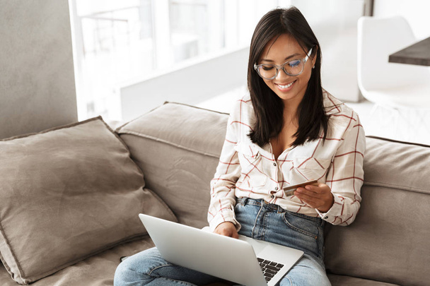 Photo of asian businesslike woman 20s holding credit card and using laptop while sitting at sofa in apartment - 写真・画像
