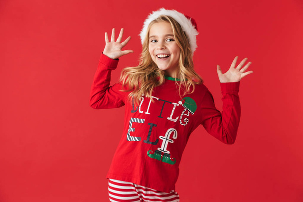 Веселая маленькая девочка в рождественском костюме стоит изолированно на красном фоне, празднуя
 - Фото, изображение