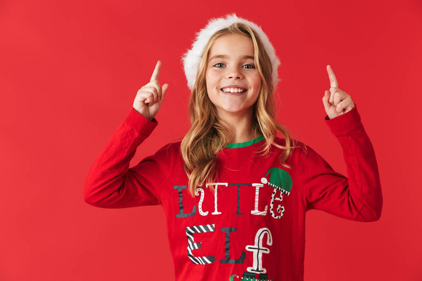 Cheerful little girl wearing Christmas costume standing isolated over red background, pointing up - Фото, зображення