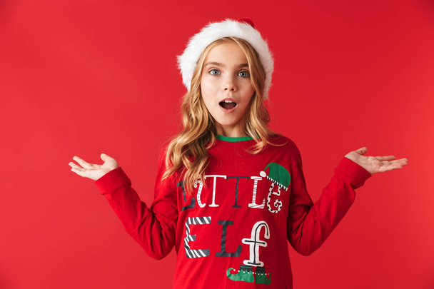 Cheerful little girl wearing Christmas hat standing isolated over red background, presenting copy space - Photo, Image